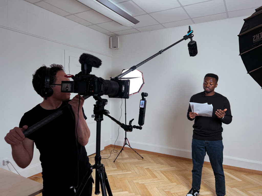 A man holding a microphone over another man's head while he reads a script aloud in front of a camera.